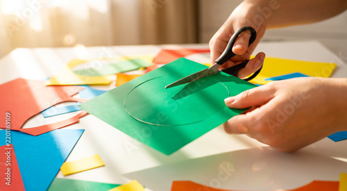 Close up of hands cutting a green circle out of paper with scissors, colorful scraps on white table, bright sunlight. Crafting, DIY hobby, creative arts, education.