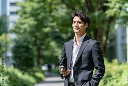 Obraz na płótnie 都会の緑道でスマートフォンを持ち自信満々に歩む日本人男性