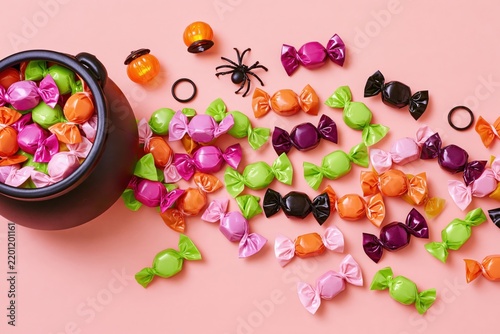 Colorful halloween candy spilling from black cauldron on pink background, including wrapped sweets, plastic spider and pumpkin decorations
