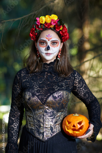 Halloween beauty portrait of woman in forest with Halloween pumpkin in hand. Santa Muerte makeup.