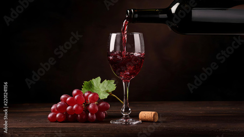 Red wine is poured from a dark glass bottle into a clear wine glass, with a bunch of red grapes and a cork resting on a dark wooden surface.