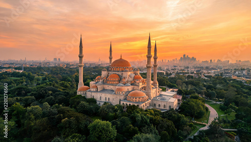 A grand historic mosque with multiple domes and minarets stands amidst greenery, with a city skyline at sunset in the background.
