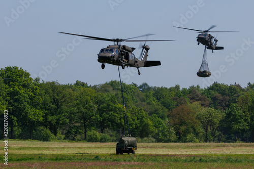 Two UH-60 Black Hawk helicopters carry a military vehicle and trailer via external sling load during a training exercise in a forested landscape.