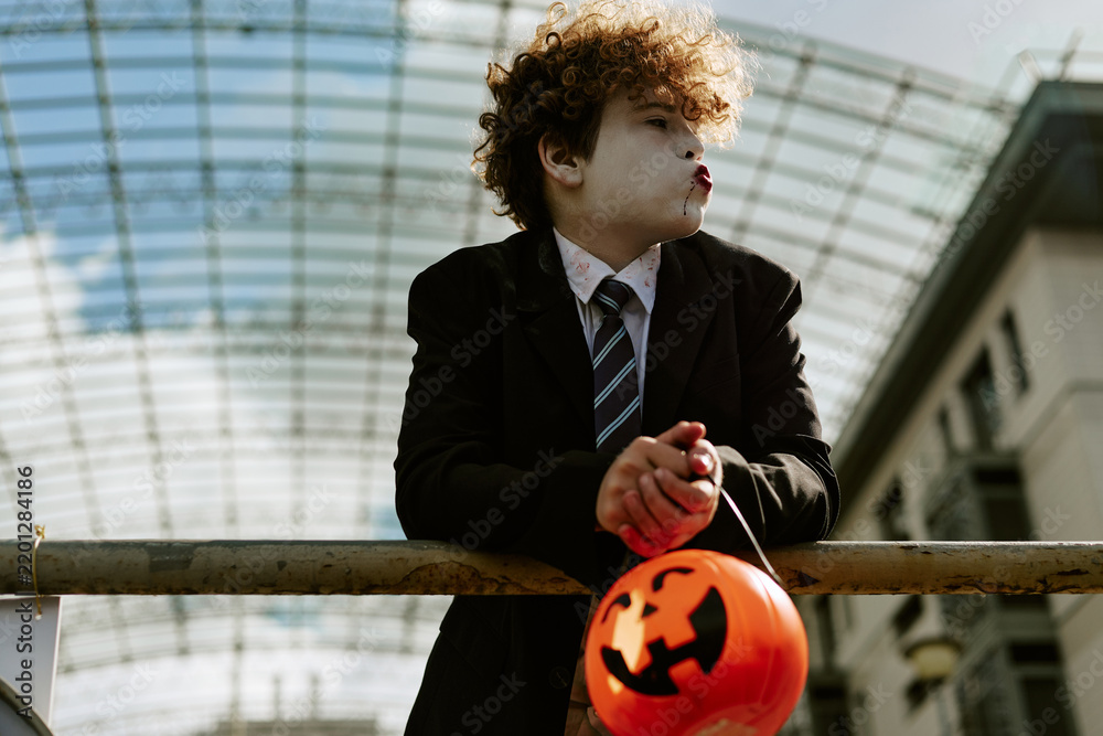 Fototapeta premium Caucasian child wearing Halloween costume makeup leaning on railing outdoors holding pumpkin bucket. Image suitable for seasonal holiday marketing and trick or treat event promotion