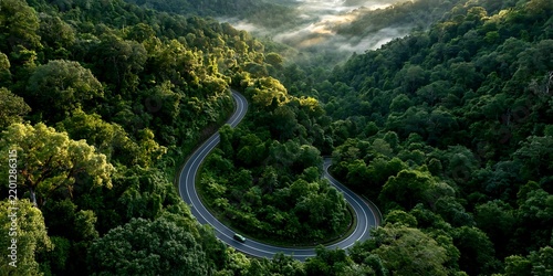 Aerial drone view of serpentine curved asphalt mountain highway through lush green misty tropical rainforest canopy with single vehicle driving at morning sunrise landscape