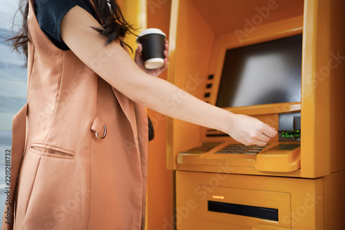 Young Asian Woman Using ATM Machine While Holding Coffee Cup, Smiling at Bank Terminal