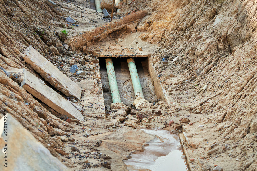 Fototapeta premium Deep excavated trench reveals two green pipes running through concrete casing at construction site during municipal utility repair project. Excavated trench, exposed pipes laid in concrete channel