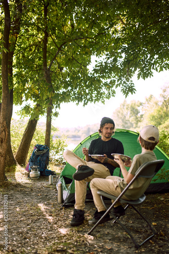 Father and son eat from metal plates on camp chairs beside tent. Family meal, outdoor living and camping comfort concept.