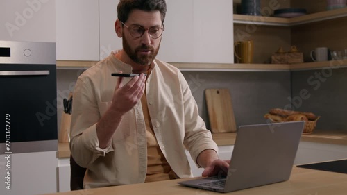Multitasking Caucasian business man at kitchen working laptop recording voice message mobile phone remote focused busy freelancer in glasses businessman guy male speaking telephone AI app indoors