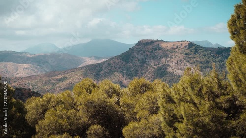 Layered rugged mountain ridges and deep valleys stretching under a partly cloudy sunlit sky framed by pine trees in El Chorro Andalusia Spain showcasing a vast Mediterranean panorama