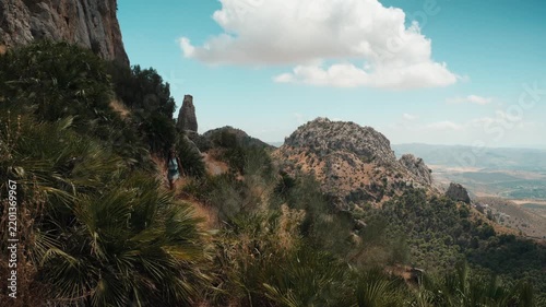 A solo female hiker walking a narrow dirt path toward the camera through dramatic limestone peaks and scrub vegetation under a bright blue sky in El Chorro Andalusia Spain
