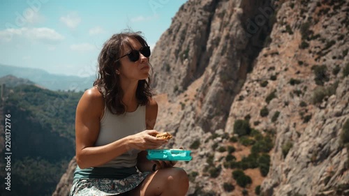 A woman sitting on a rocky ledge eating lunch while looking out over a deep mountain gorge in El Chorro Andalusia Spain in bright natural light on a solo hiking break