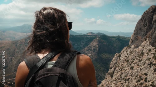 A young woman with a backpack standing on a rocky mountain overlook looking out over the vast landscape of El Chorro Andalusia Spain under a partly cloudy sky