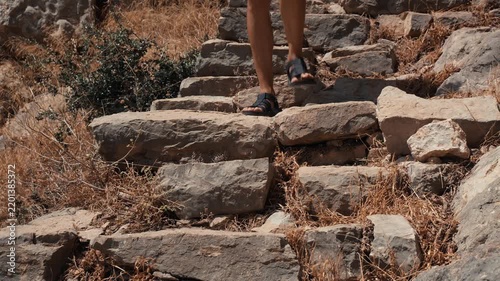 A solo female traveler climbing a weathered stone staircase surrounded by dry grass and rocks in El Chorro Andalusia Spain under bright natural daylight on an ancient mountain path