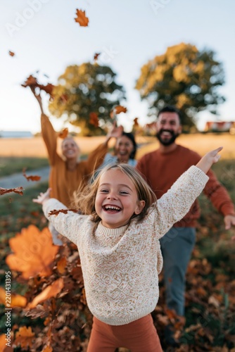Joyful child in sweater running toward camera while family throws autumn leaves in a golden field on a sunny fall day