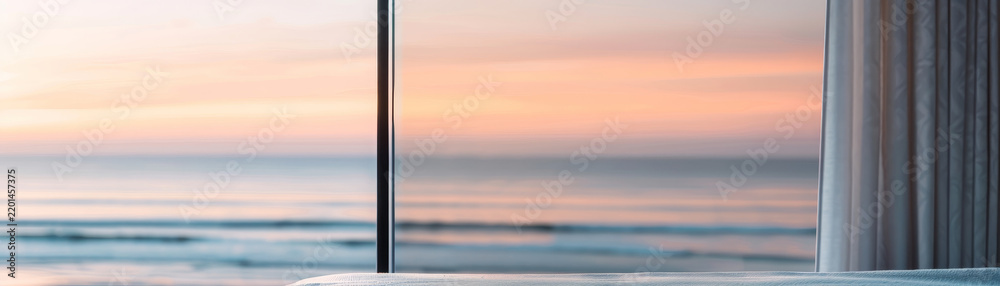 Fototapeta premium A bedroom window opens to a breathtaking view of the ocean at sunset, where gentle waves crash against the shore, reflecting the golden hues of the evening sky in a tranquil coastal setting.