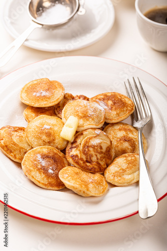Small Dutch poffertjes pancakes are piled on a white plate with red trim, dusted with powdered sugar and topped with butter, beside a fork, coffee cup, and sugar bowl
