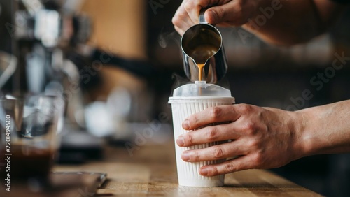 Barista pouring fresh hot espresso into a white paper takeaway cup on wooden countertop with displays close up person pouring hot coffee metal pitcher disposable paper cup