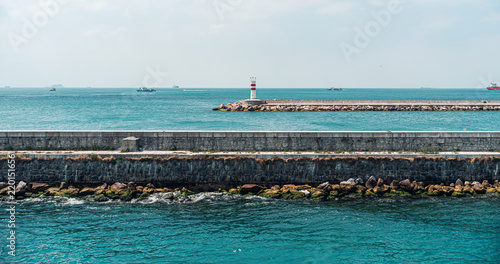 A red and white lighthouse stands on a stone breakwater overlooking the calm sea. Boats and ships are visible in the distance, enhancing the maritime atmosphere.