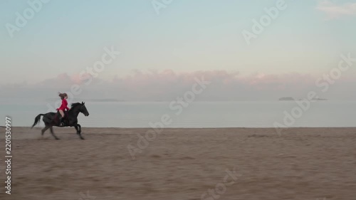 A young girl with long hair in a short red dress rides a horse on the beach at dawn. Shooting from a medium distance. Concept of a free woman rejecting imposed values.