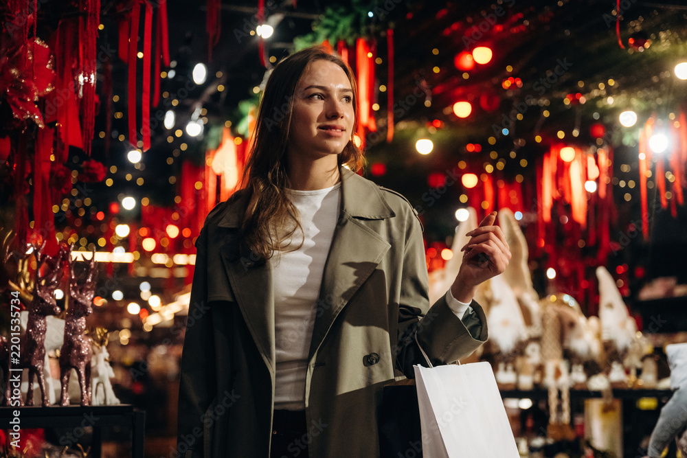 Fototapeta premium Woman carrying shopping bag while browsing brightly decorated festive market with red ornaments, warm lights, and displays in an indoor holiday shopping setting