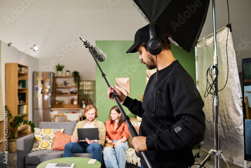 Young adult Caucasian sound technician holding boom microphone while filming young adult actors indoors. Suitable for video production, broadcasting, media industry advertising