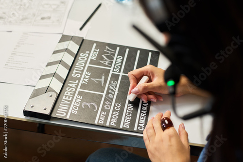 Young adult woman writing scene details on movie clapperboard during film production. Concept represents filmmaking industry, useful for cinema advertising and video production campaigns