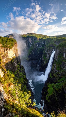 Majestic waterfall cascades down a lush green cliffside under a cloudy blue sky.