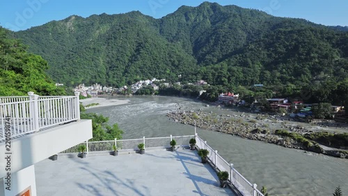 The Ganga River winds past forested hills in Rishikesh beyond a sunlit terrace with white railings.