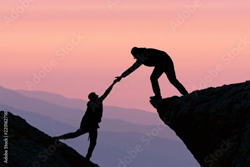 Silhouetted climbers helping each other across rocky cliff edges at sunset