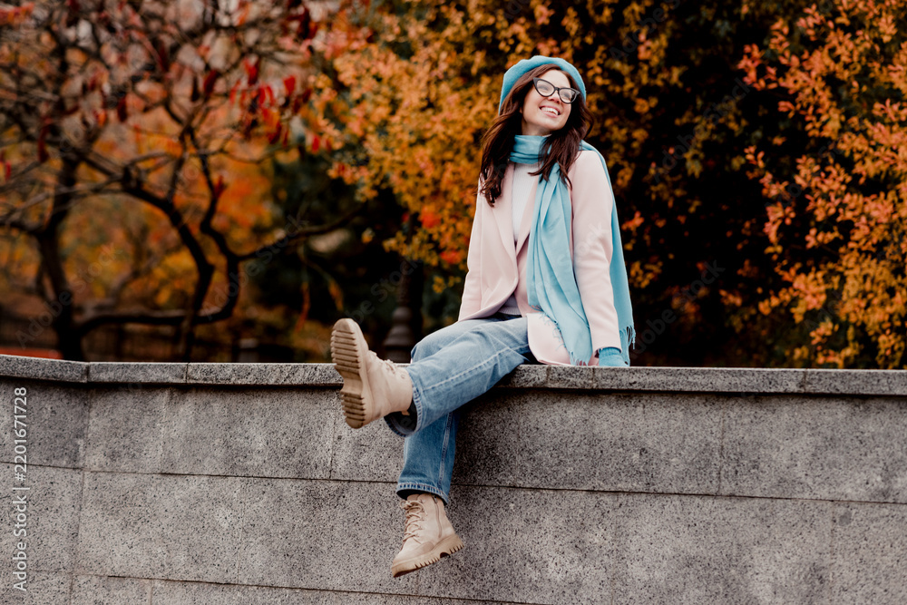 Fototapeta premium Smiling young woman sitting on a stone wall in autumn wearing a blue scarf and beret enjoying fall outdoors