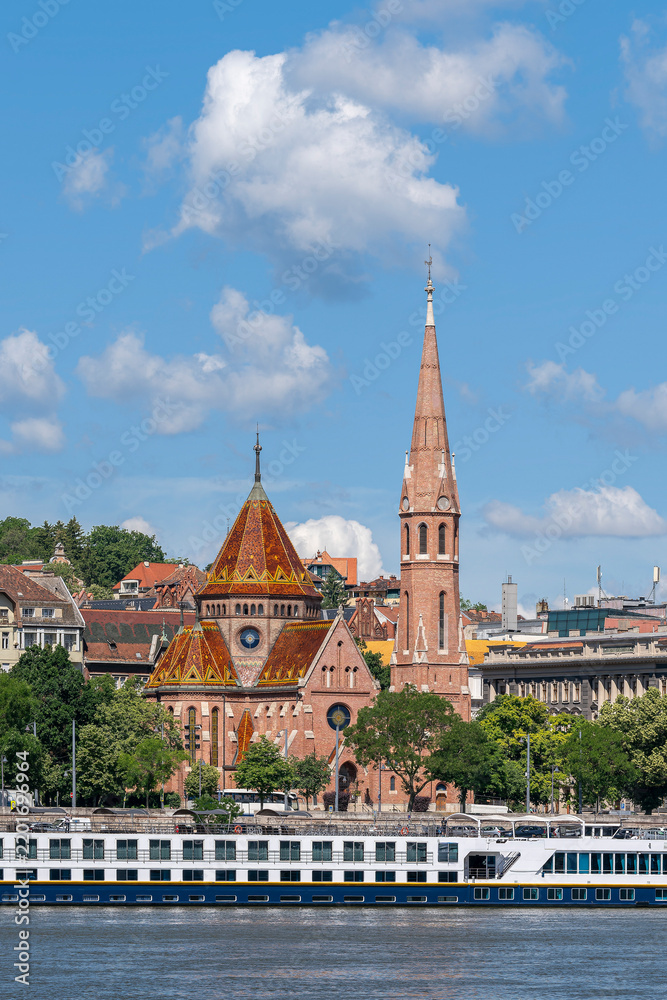 Fototapeta premium Red-brick Reformed church with a patterned tiled roof and tall spire standing beside the Danube embankment in Budapest
