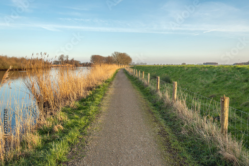Narrow road along a dike in a Dutch polder. At the foot of the dike is a fence made of wooden posts and wire mesh. The photo was taken on a slightly cloudy day at the end of the winter season.