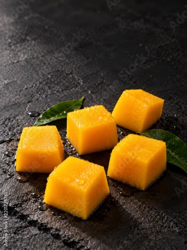 Five mango cubes on dark charcoal stone with moody side lighting and ample negative space