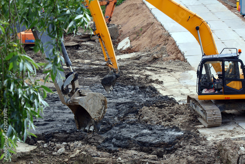 Two yellow tracked excavators operate on a muddy construction site, digging into dark, loose soil beside a paved walkway