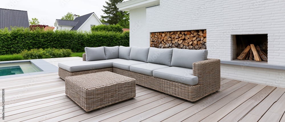 Fototapeta premium L-shaped wicker sectional sofa with grey cushions on a wooden deck beside a swimming pool and white brick fireplace in a suburban backyard
