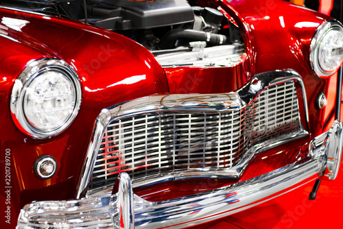 Glossy red vintage automobile front with round headlights, chrome grille, polished bumper, and open hood displayed as a classic car closeup at an indoor exhibition