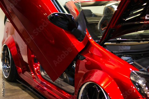 Glossy red customized sports car with raised scissor door, black mirror, polished wheels, and visible interior displayed indoors at an automotive exhibition
