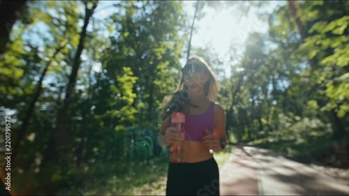 Young woman jogging along sunlit forest trail holding water bottle wearing sunglasses and sportswear