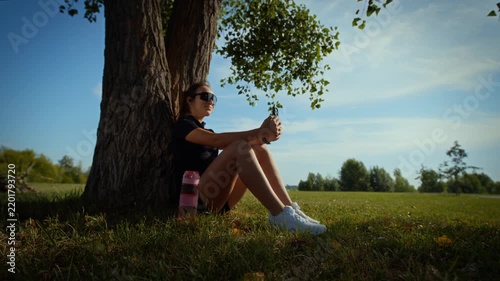 Relaxed young woman raising smartphone beneath tree in sunny park with open meadow and blue sky view