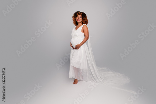 Foto Pregnant woman in white flowing maternity gown standing barefoot, smiling and cr