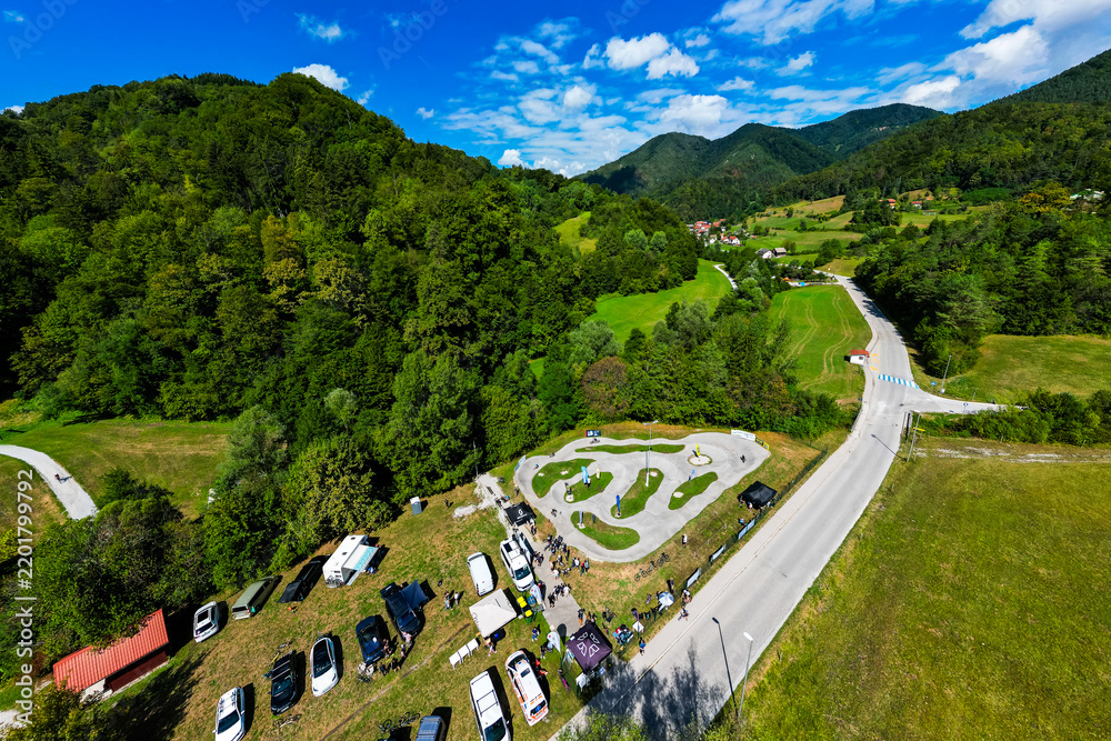 Obraz premium A wide aerial view of the asphalt pumptrack park in Zagorje ob Savi, Slovenia, surrounded by green fields, rolling hills, a railway line, and a bright blue sky filled with fluffy white clouds.