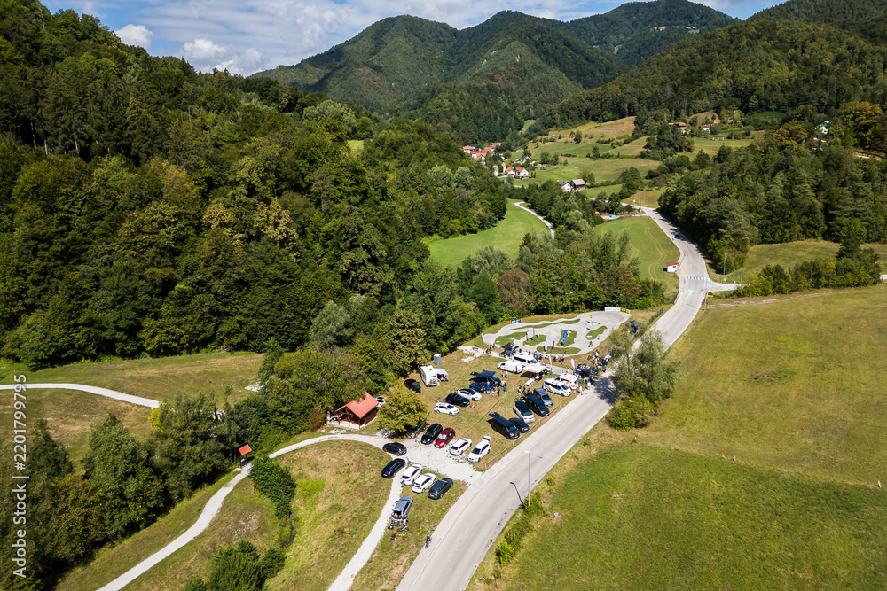 Obraz premium A wide aerial view of the asphalt pumptrack park in Zagorje ob Savi, Slovenia, surrounded by green fields, rolling hills, a railway line, and a bright blue sky filled with fluffy white clouds.