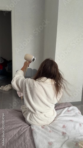 Person in a white robe drying hair while seated on a bed