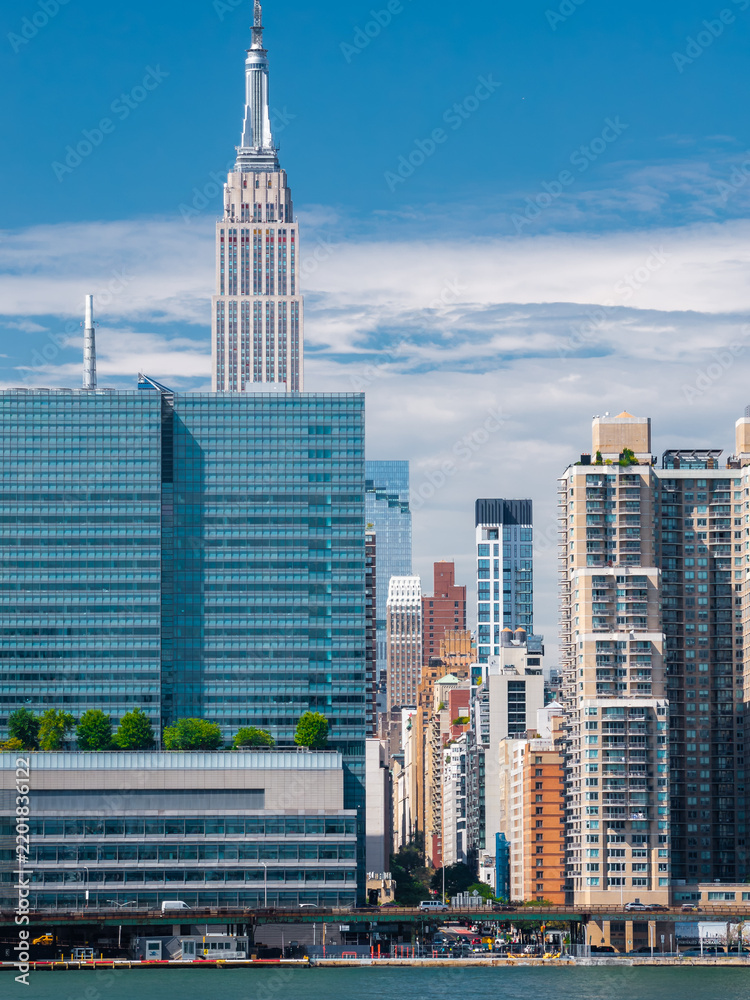 Fototapeta premium Empire State Building rises over modern waterfront glass skyscrapers and highway traffic in Manhattan, New York City on a bright summer day.