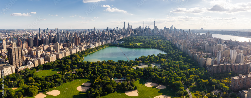 Obraz premium Aerial view displays Central Park reservoir surrounded by trees, baseball fields, and the Midtown Manhattan skyscraper cityscape under clear skies in New York City.