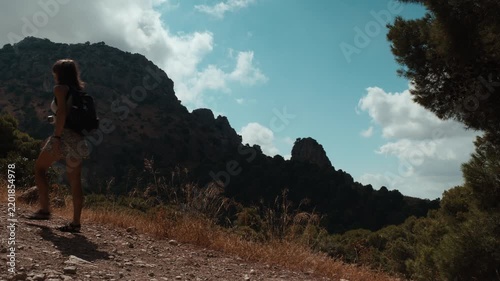 A young woman hiker walking steadily along a rugged rocky mountain trail past dry grass and open scrubland under a blue sky with scattered clouds in El Chorro Andalusia Spain
