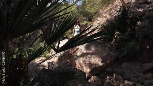 A young woman hiking along a rugged rocky trail surrounded by fan palms and large boulders in a sun dappled Mediterranean forest in El Chorro Andalusia Spain