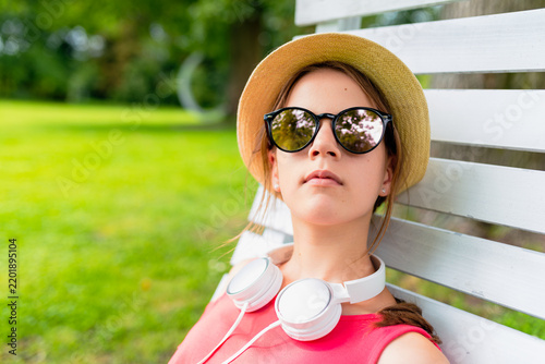 Close up portrait of a relaxed teenage girl wearing a straw hat, sunglasses, and white headphones, sitting on a white wooden chair in a sunny green summer park.