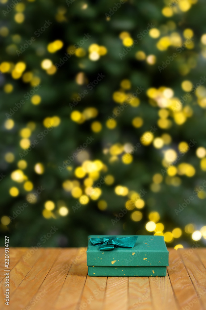 Obraz premium Green Christmas gift box with decorative bow on wooden surface against blurred Christmas tree and warm golden bokeh lights